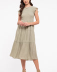 Brunette model wearing light olive colored dress, features a short ruffle sleeve, fitted smocked top, and flowy skirt with 3 tiers. Dress hits her at mid-calf. She wears with brown ankle boots.
