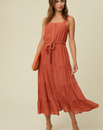 Model wearing a rust (red-orange) dress, features buttons all the way down the front, a drawstring at waist, sleeveless with a square neckline. Skirt has flowy tiers. Model paired with sandals and a bright woven clutch.