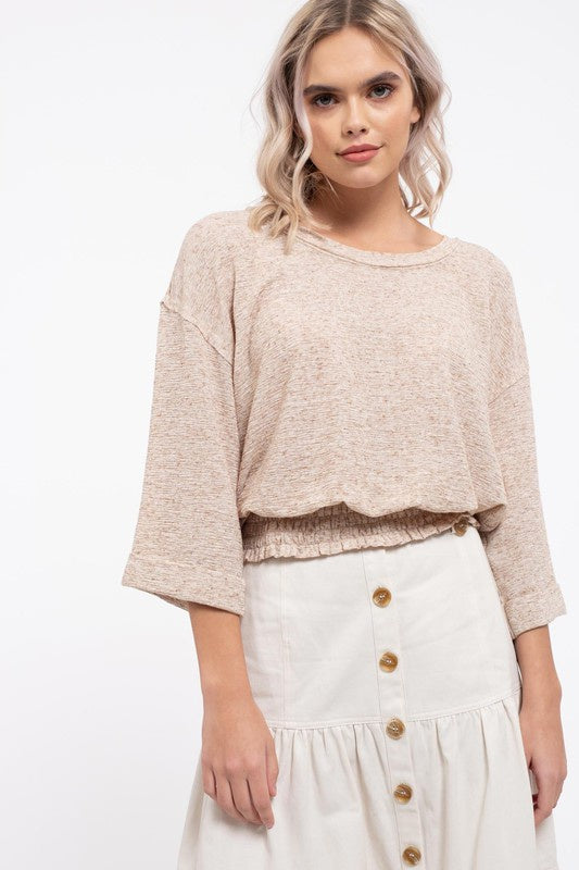 Blond Haired model wears the two tone top, shows wide sleeves, dropped shoulder, round neckline and smocked bottom hem giving the shirt more volume. Paired with an off white button front skirt.