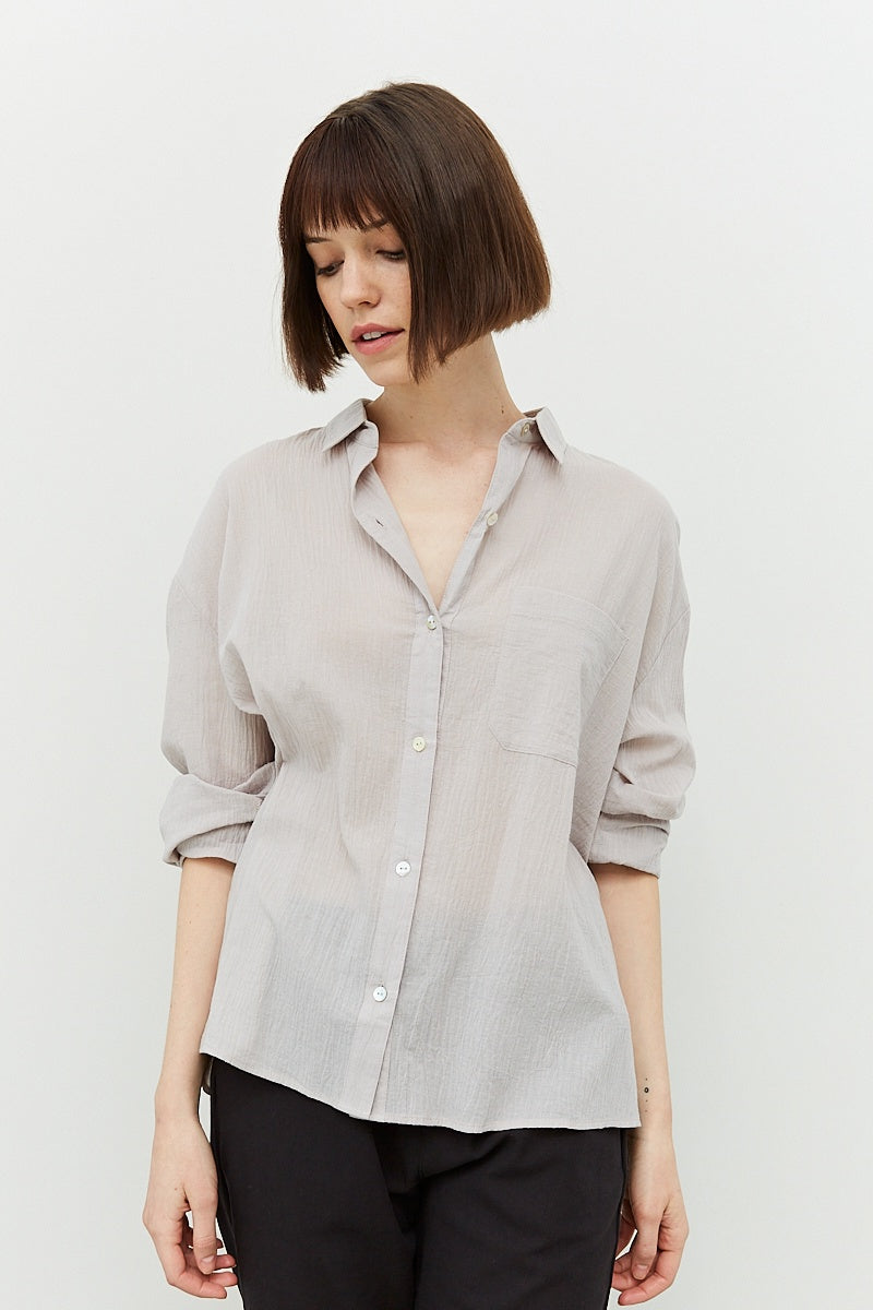 model with a brown bob wearing the Marta shirt, 3/4 buttoned with sleeves rolled up. Shows the straight hem and txtured weave of the lgihtweight material. Color is a very pale grey with undertones of purple.