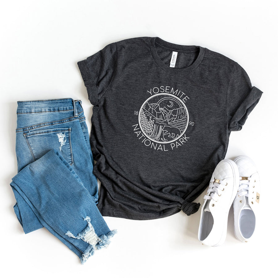 Flat lay of knotted Charcoal grey Yosemite t-shir, shirt has white illustration of mountains, a moon, and a bear. Paired with distressed denim and white sneakers.