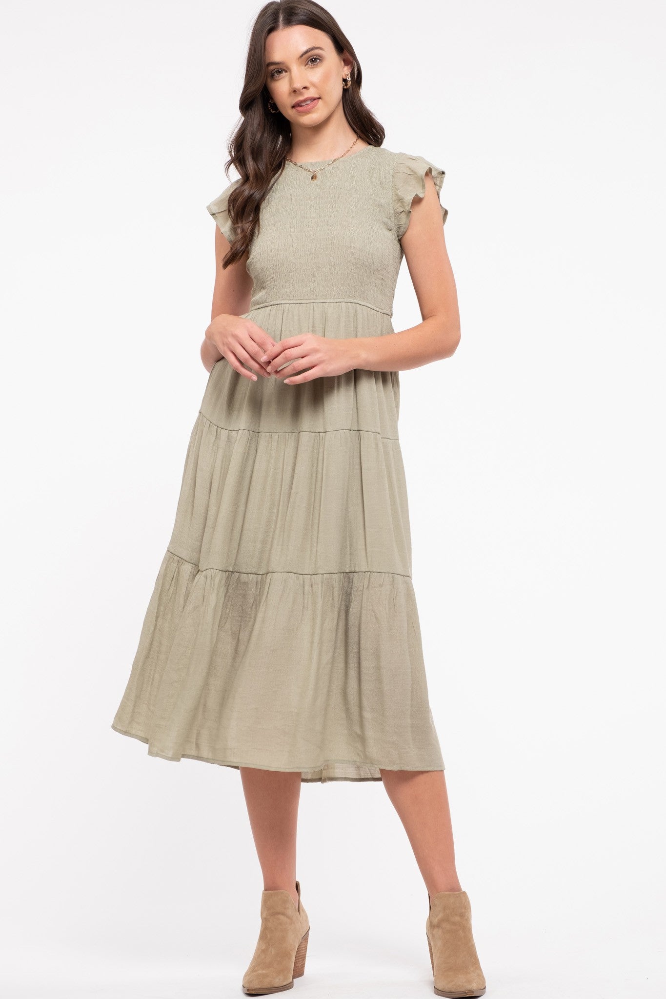 Brunette model wearing light olive colored dress, features a short ruffle sleeve, fitted smocked top, and flowy skirt with 3 tiers. Dress hits her at mid-calf. She wears with brown ankle boots.