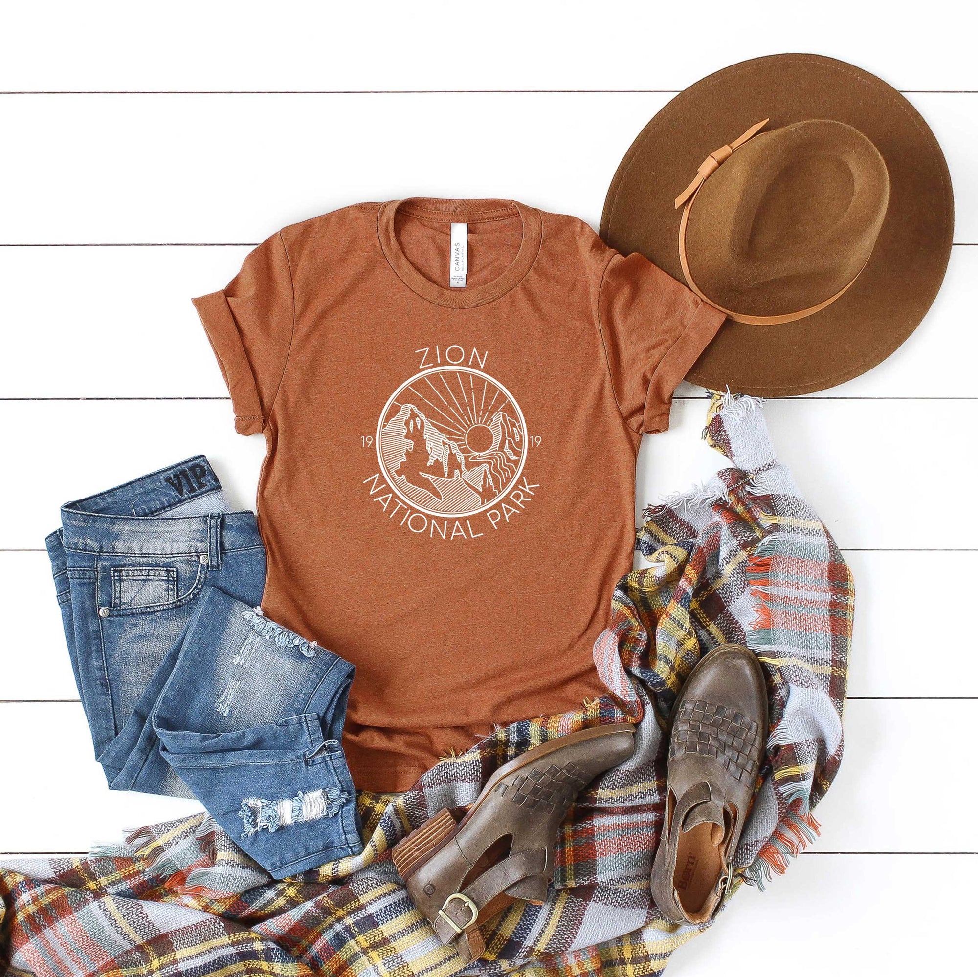 Burnt orange tee with cuffs rolled, features white ink design of Zion National Park with a sunrising over the moutnains in a circle design. Flat lay shows the shirt paired with a large brim brown hat, distressed denim and buckle ankle boots