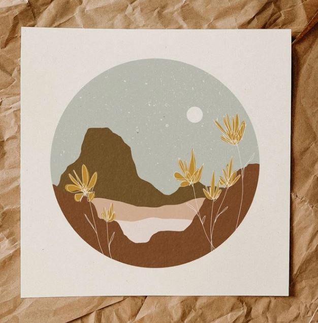 An evening desertscape. with mustard yellow flowers in the foreground, taup, brown and peach mountains behind them and a pale blue, speckled sky and white moon. Image is in a circle shape with white border on square print